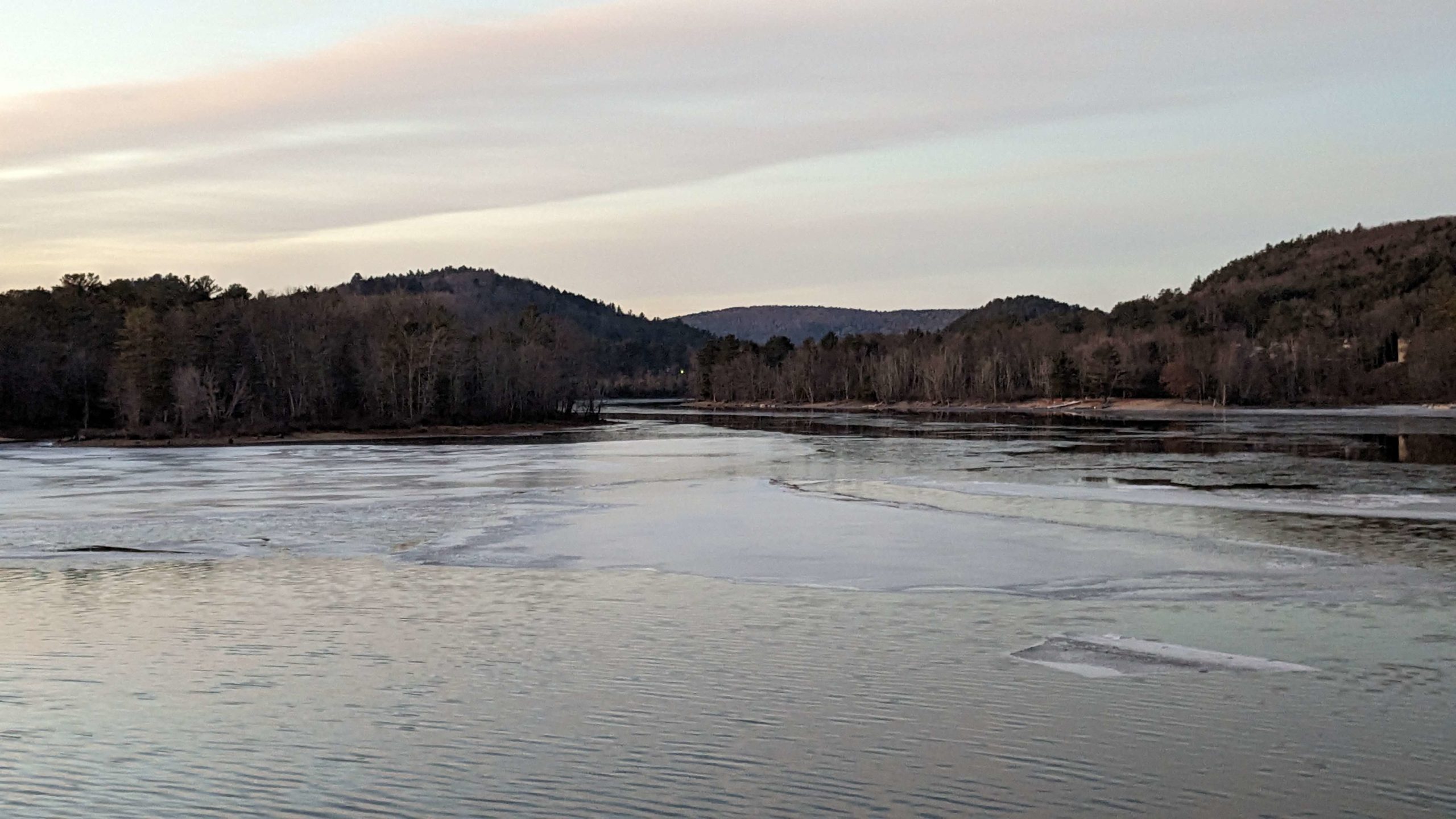 A lake with sheets of ice on top, with mountains in the horizon. The sky is mostly covered in a thin, light-grey cloud with blue sky peeking through the cracks.